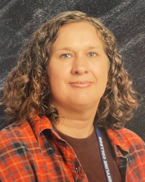 Woman with black hair, wearing light blue shirt, smiling, key necklace, earrings. Background: gray.