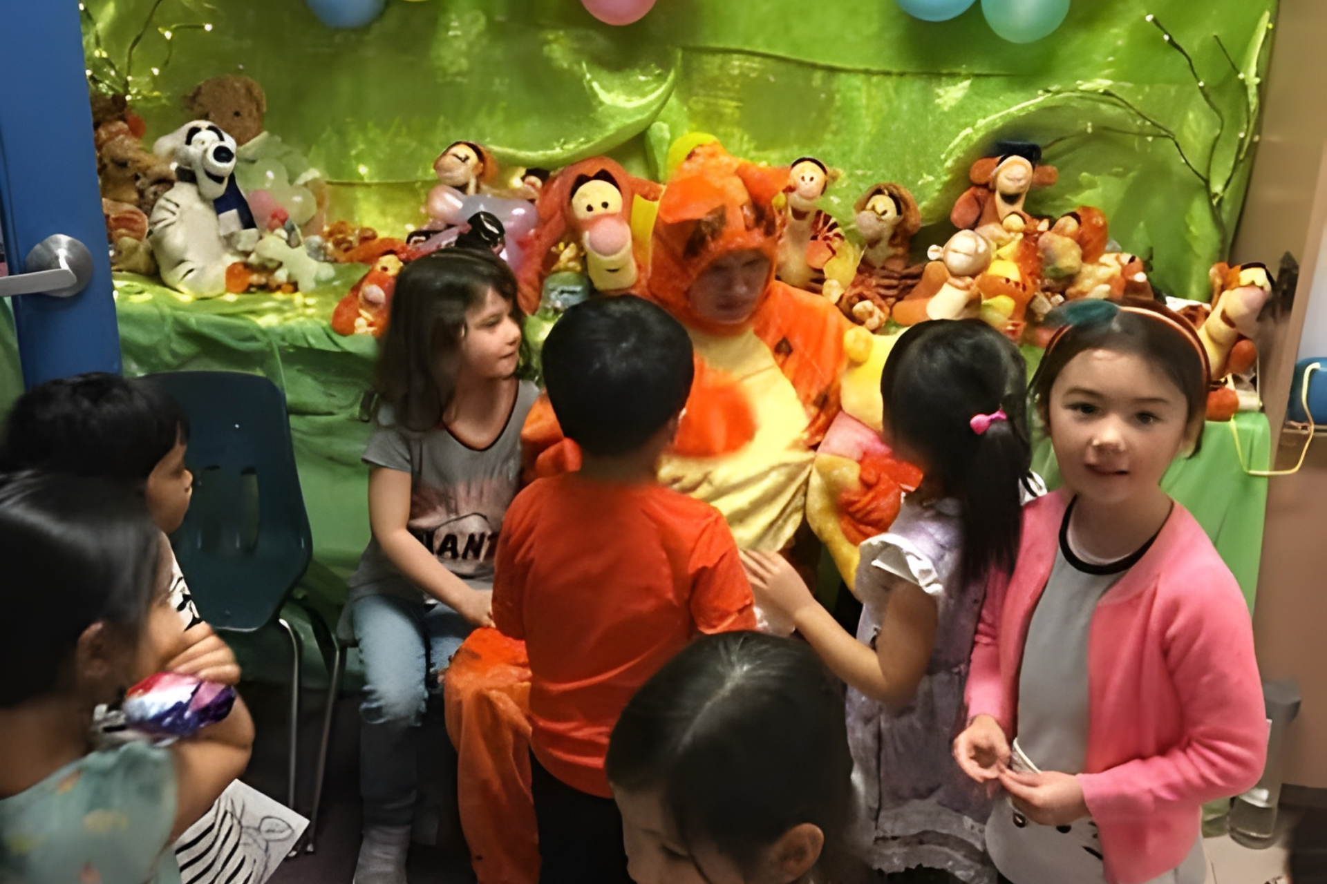 Children gather around a person in a Tigger costume, surrounded by stuffed animals and decorations.