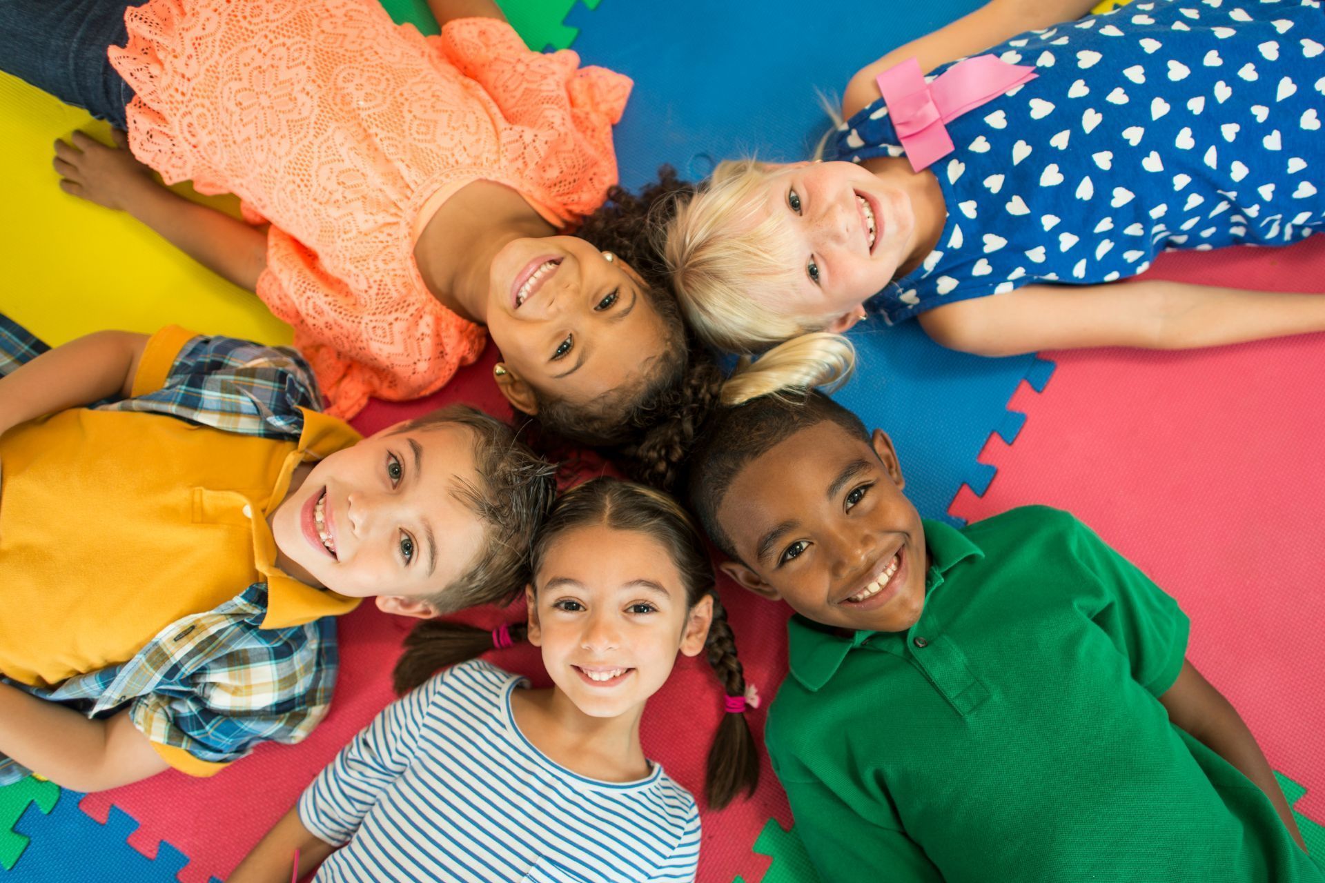 Group of smiling children lying on colorful mats in a circle, looking up.