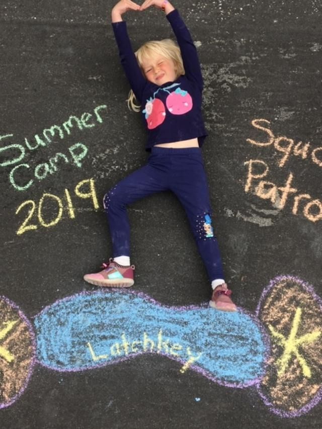 Child in blue outfit making a heart shape with arms, standing on a chalk drawing that says 