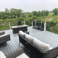 Outdoor deck with gray composite flooring, black railing, modern furniture, fire pit, and a view of greenery.