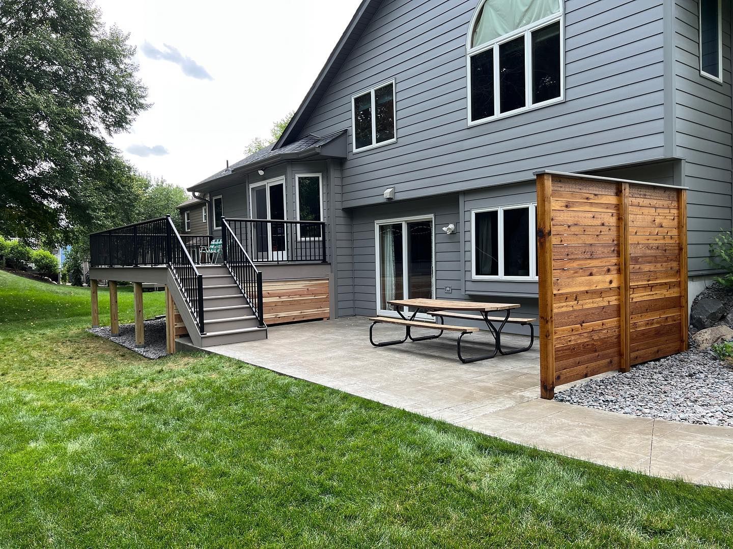 Backyard patio with wooden privacy screen, picnic table, and deck beside a gray house.