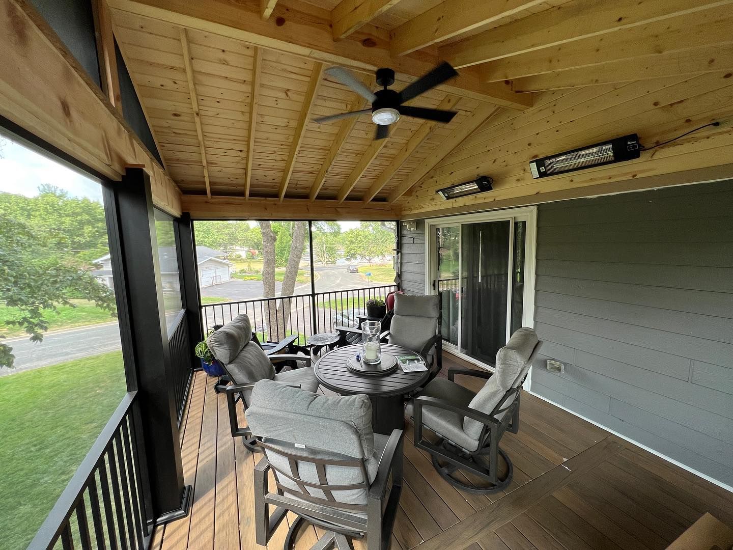 Screened porch with gray furniture, ceiling fan, and heating elements. View of a yard.
