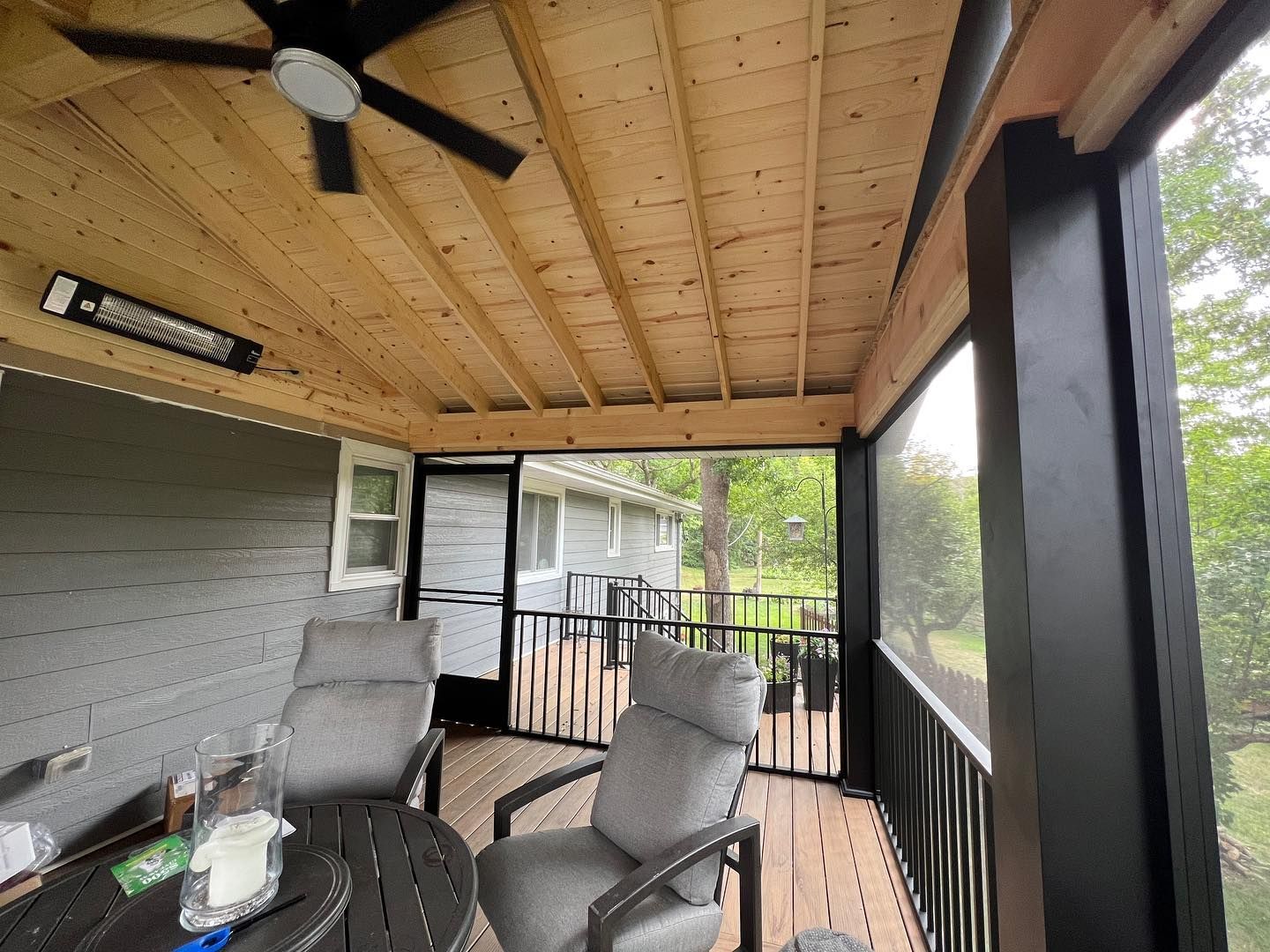 Screened porch with gray chairs, black railing, wooden ceiling, and an infrared heater.