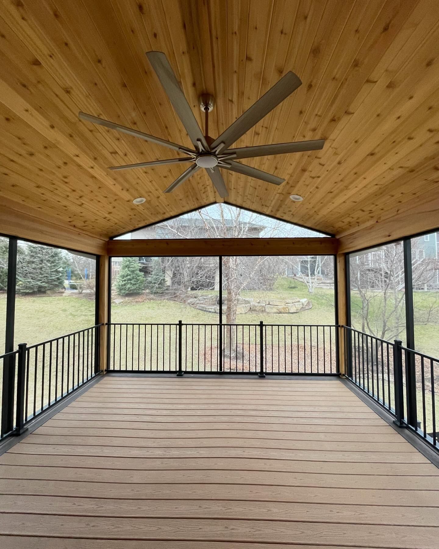 Screened-in porch with wood ceiling and floor, black railing, large ceiling fan, and view of trees.