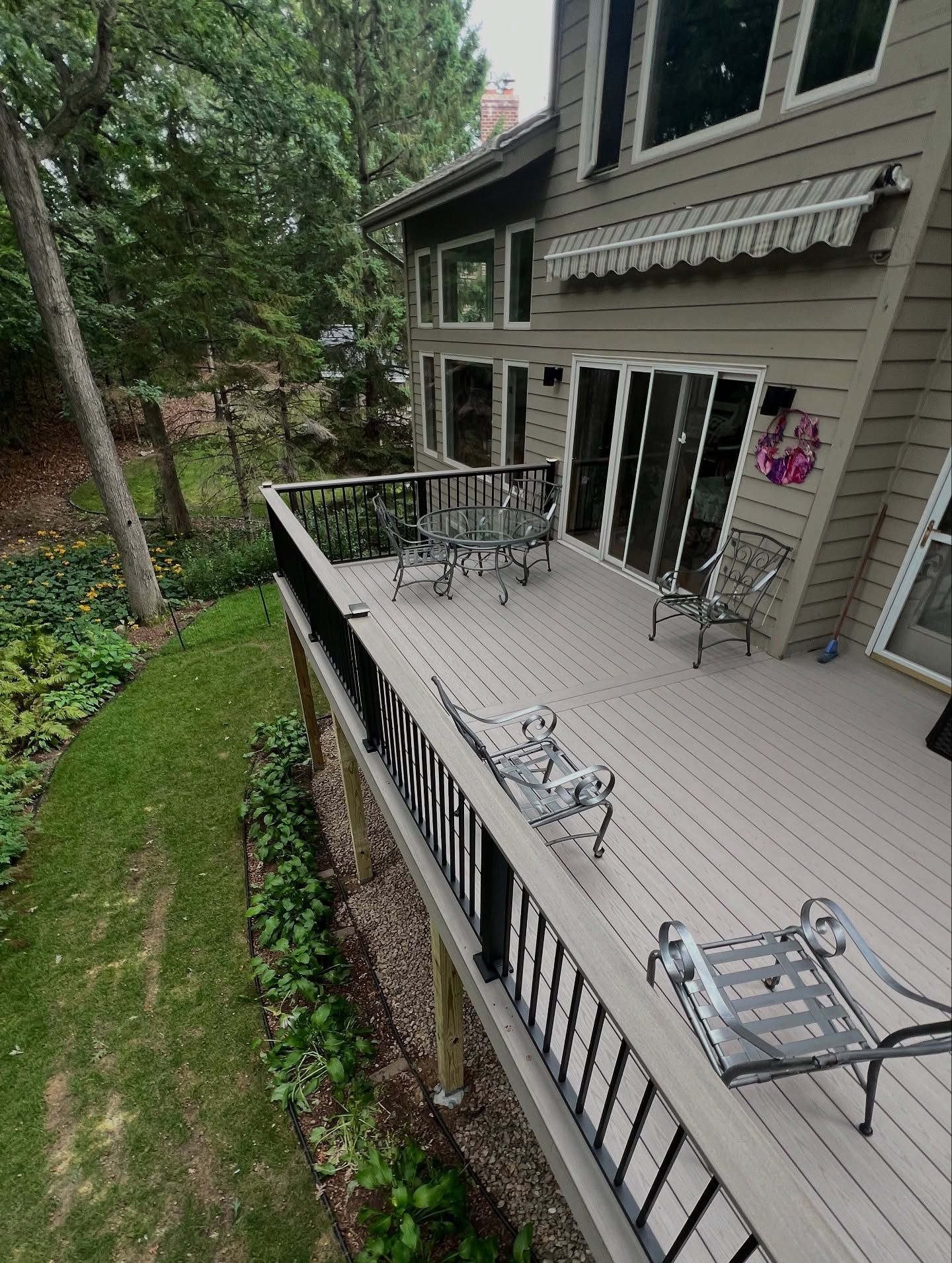 Wooden deck with metal furniture overlooking a grassy yard with trees. House has sliding doors.