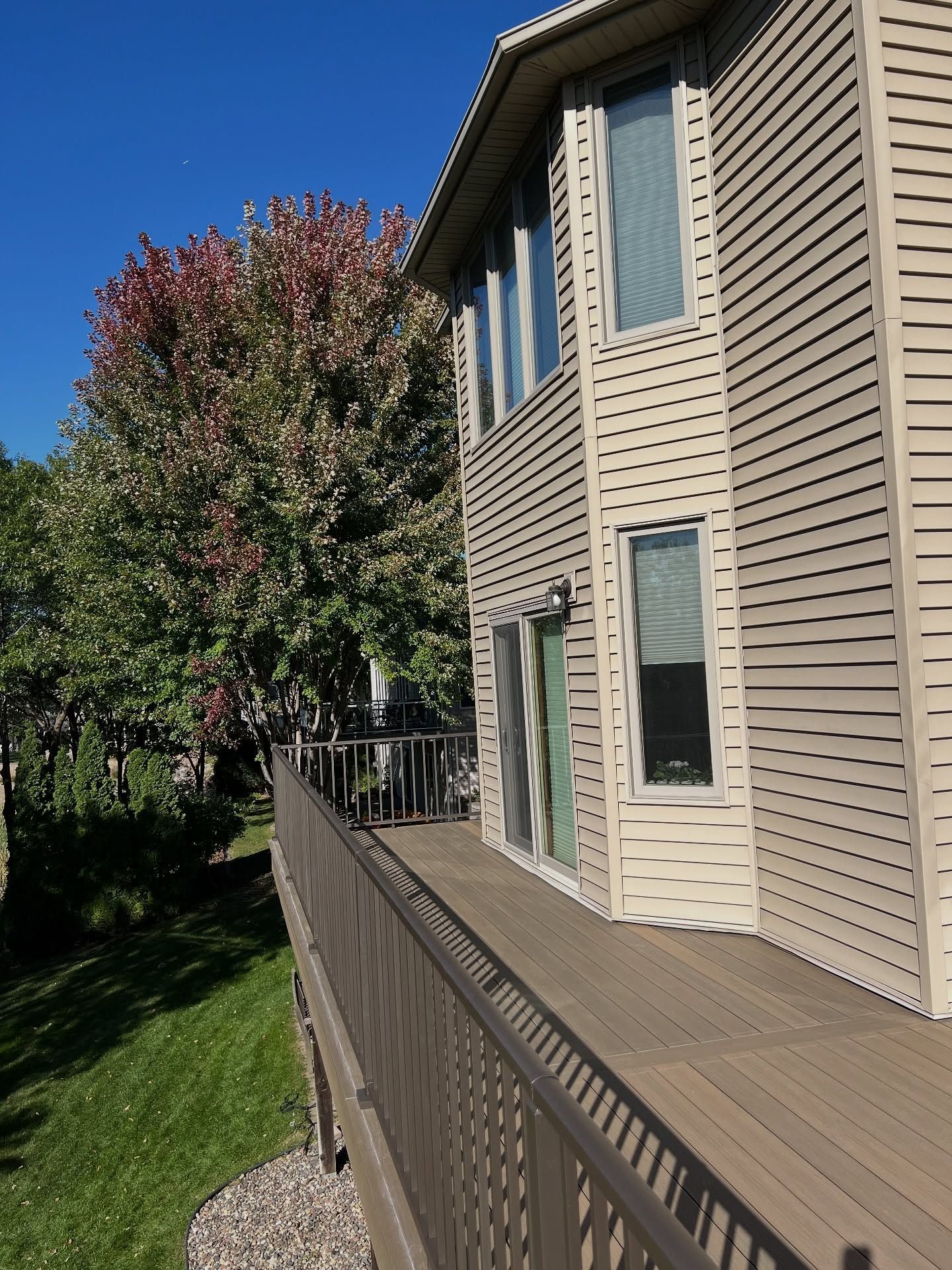House with a brown deck and siding, next to a grassy hill and trees with red and green leaves under a blue sky.