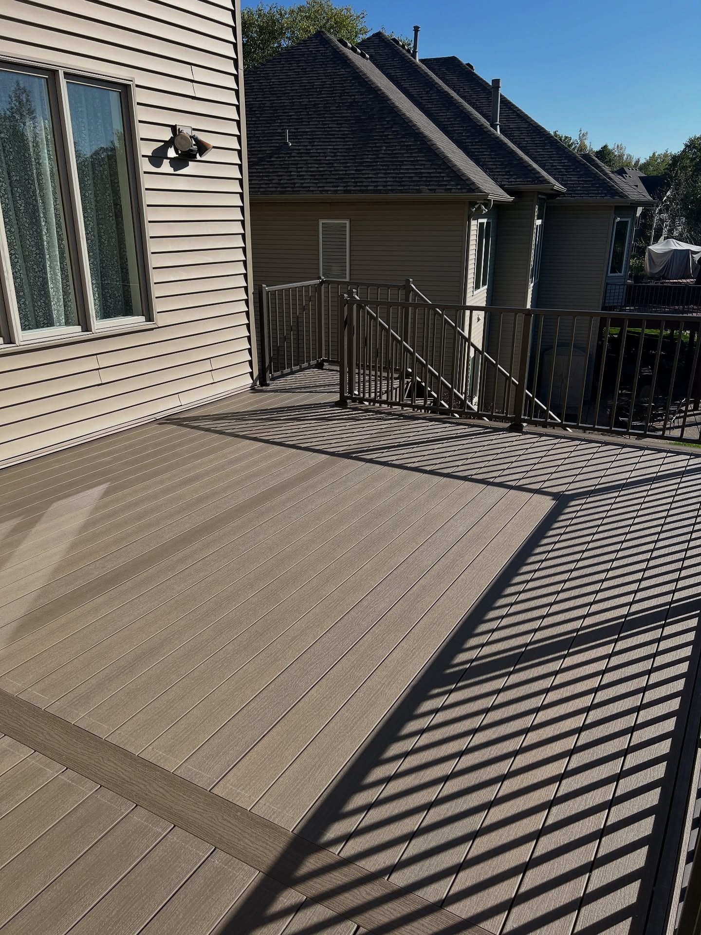 Wooden deck with railings, shadow cast from fence, next to a beige house with windows.