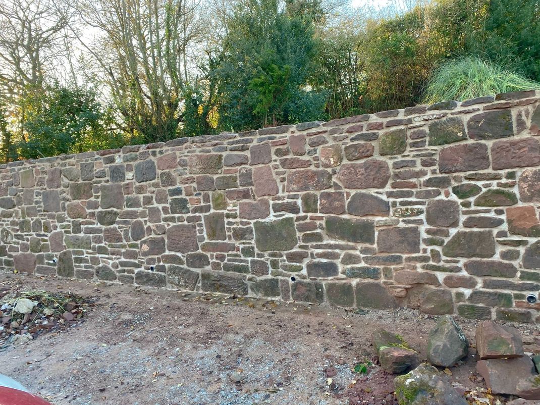 A large stone wall with lime mortar, is surrounded by trees and gravel.
