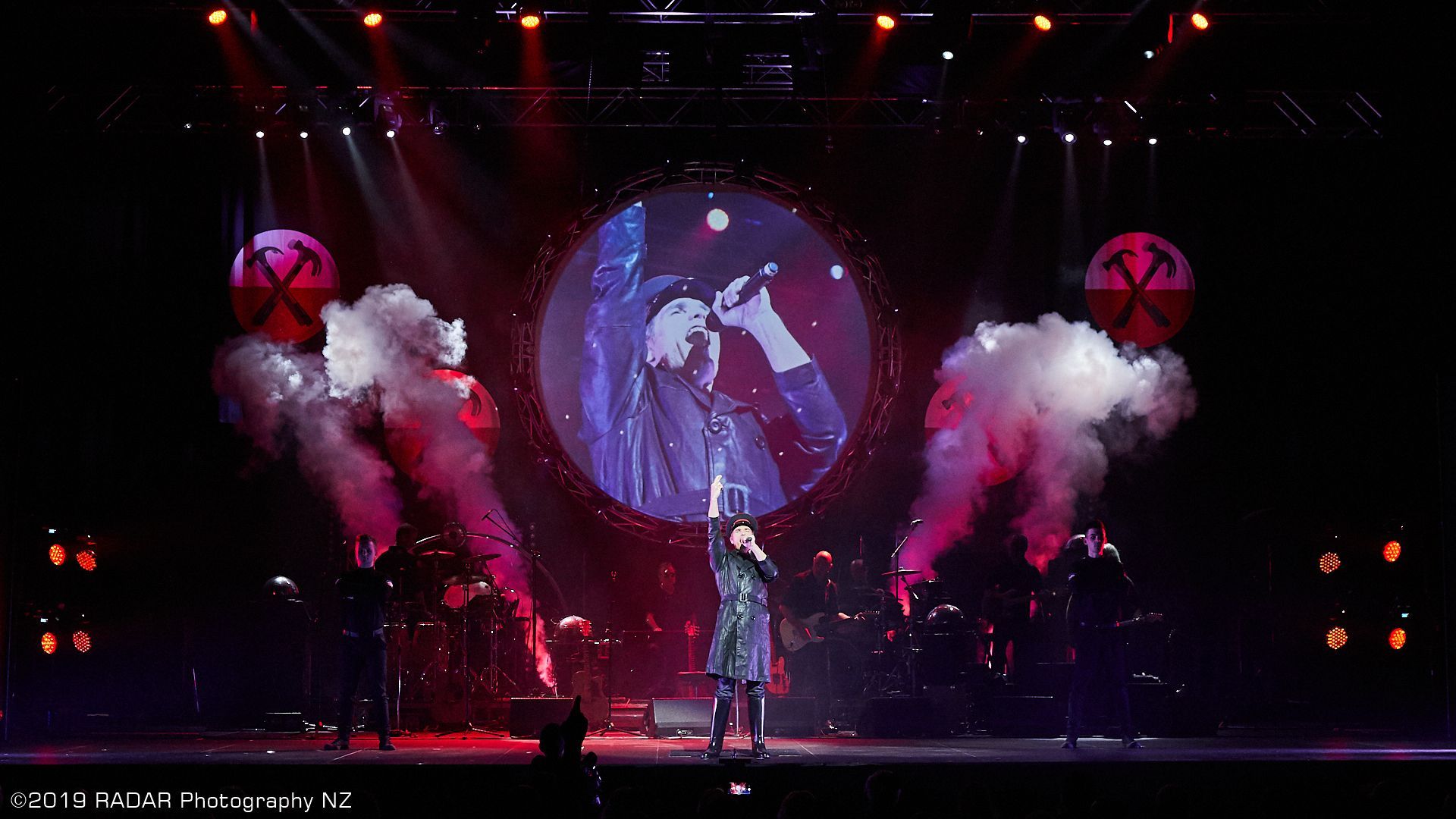 A singer performs on a stage framed by two red logo globes, plumes of smoke, and a large circular screen backdrop.