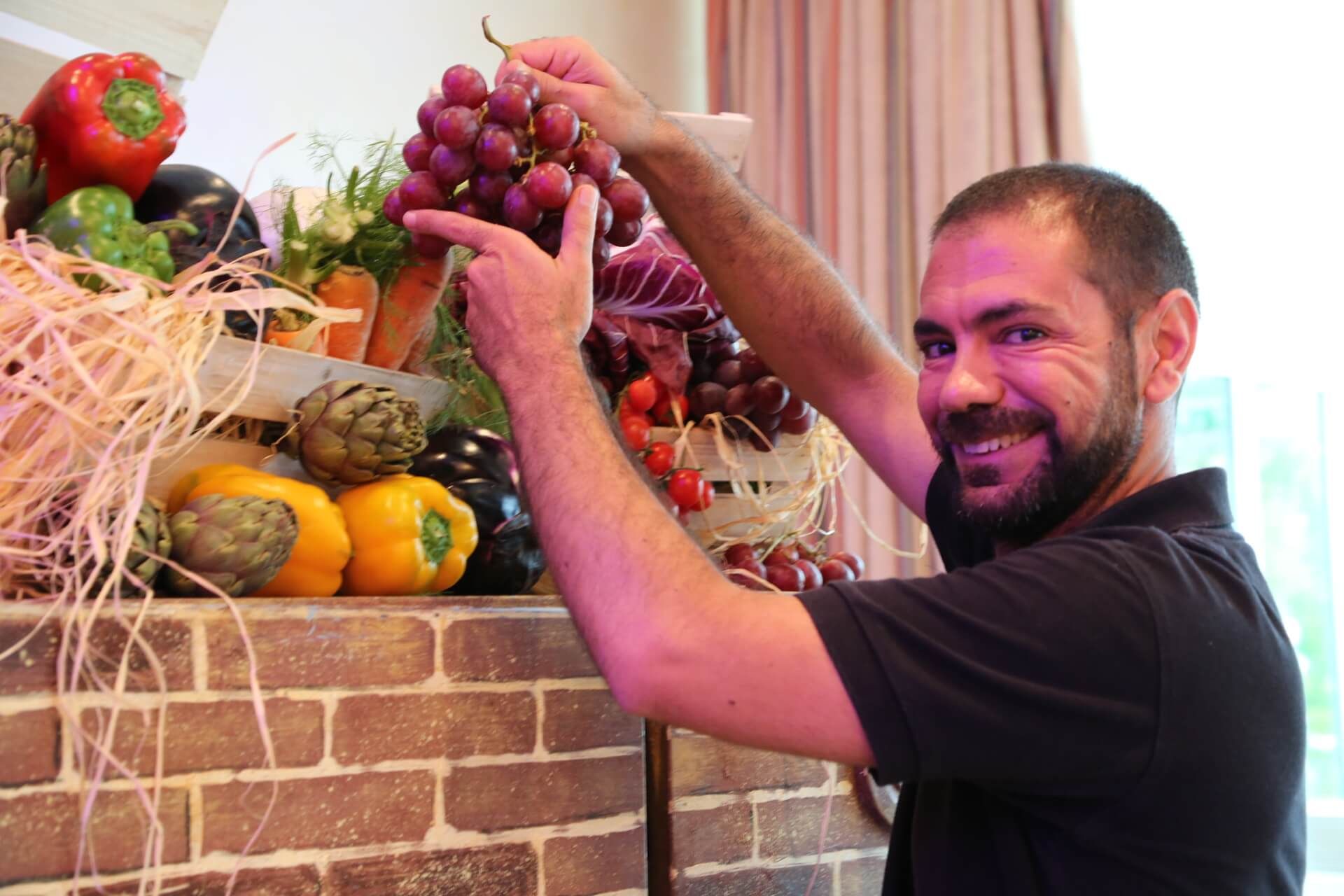 Man pointing to grapes; produce display of vegetables, fruit, on brick shelf.
