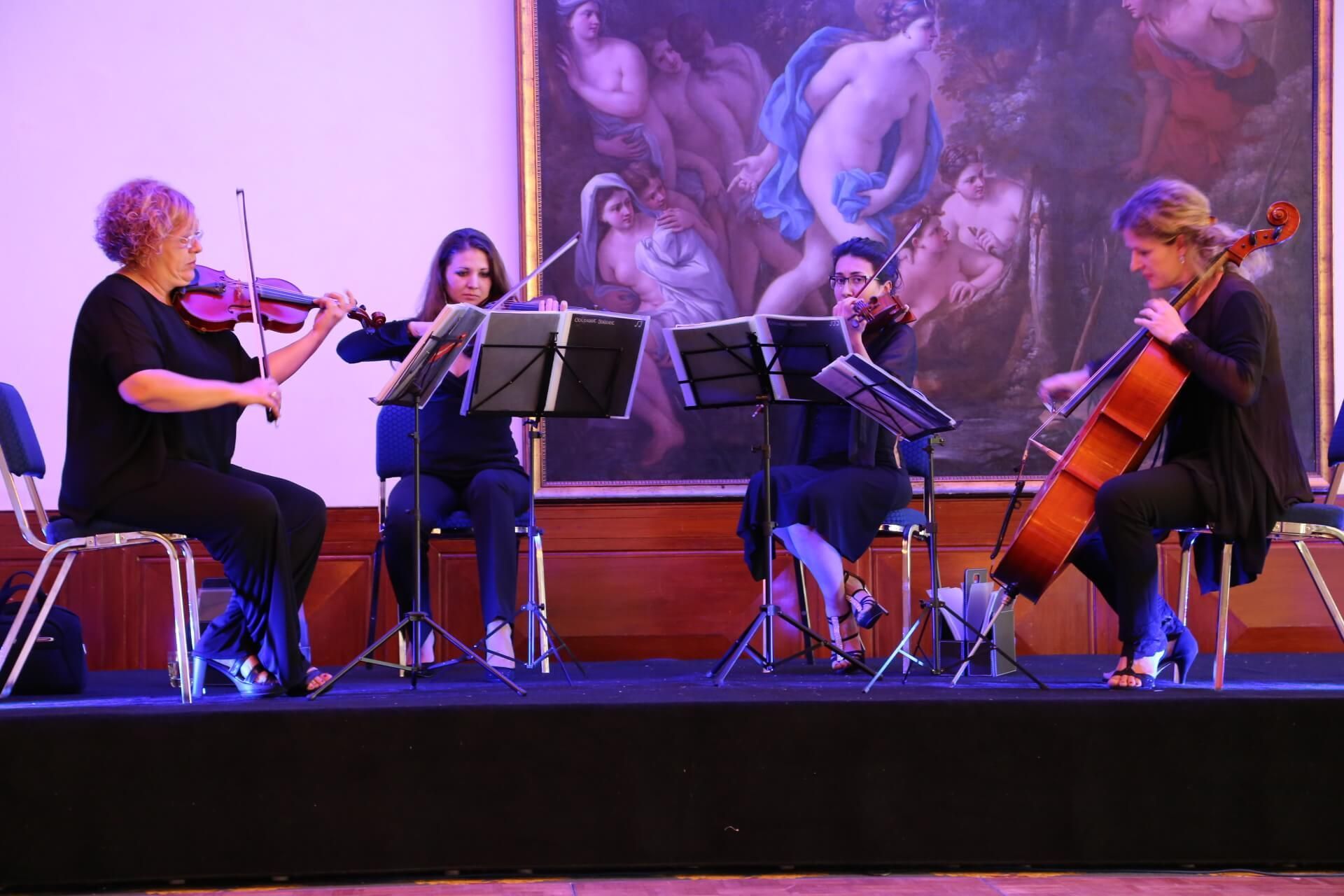 String quartet performing on stage in front of a painting. Musicians playing violin and cello, with music stands.