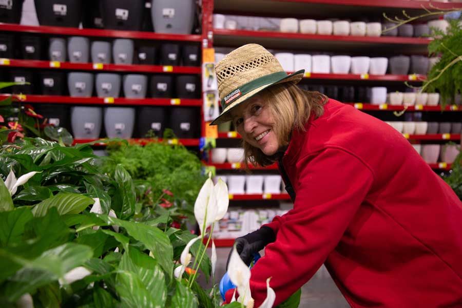 Woman in a hat and red jacket examines plants in a store, smiling. Pots on shelves in the background.