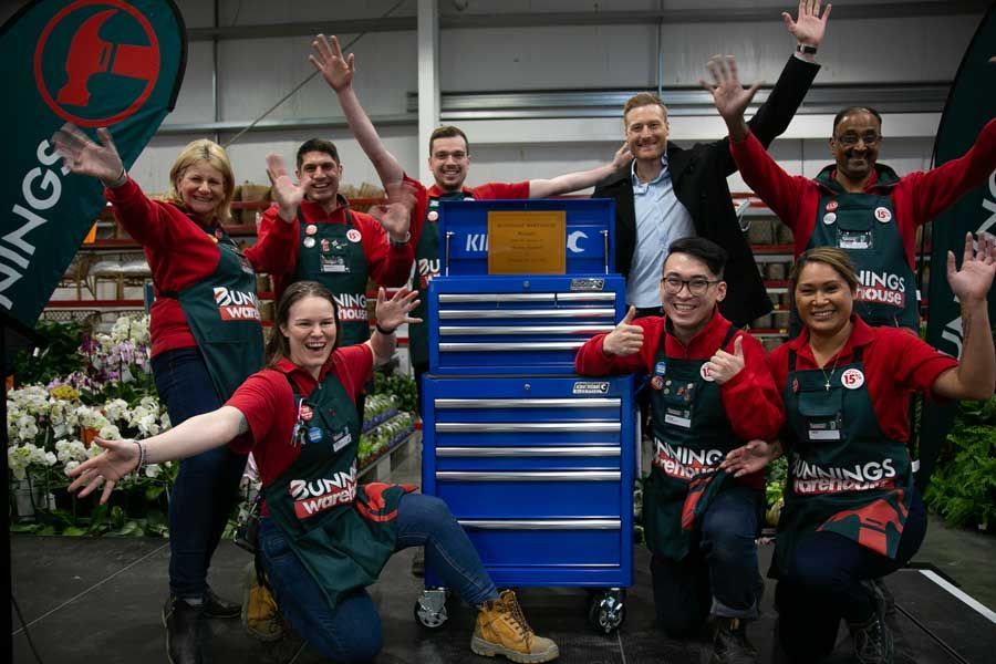 People in red shirts and aprons with a blue tool chest celebrating inside a store.