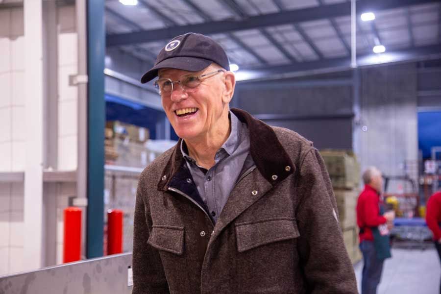 Smiling man in a cap and jacket, indoors, appearing to laugh, in a warehouse setting.
