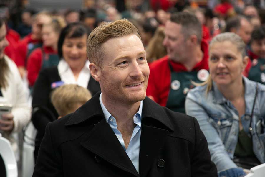 Man with red hair smiles, wearing a black coat and blue shirt, seated among a crowd of people.