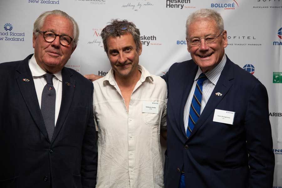 Three men pose at an event; one in a light shirt, two in suits. All are smiling, shoulder-to-shoulder.