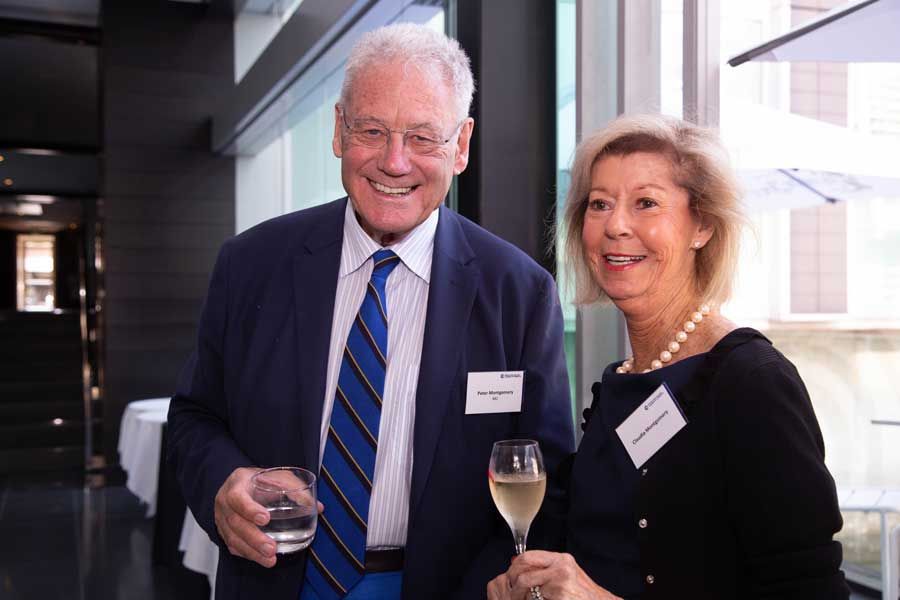 Man and woman smiling, at an event. Man in a blue blazer holds a drink. Woman in black top holds a glass.