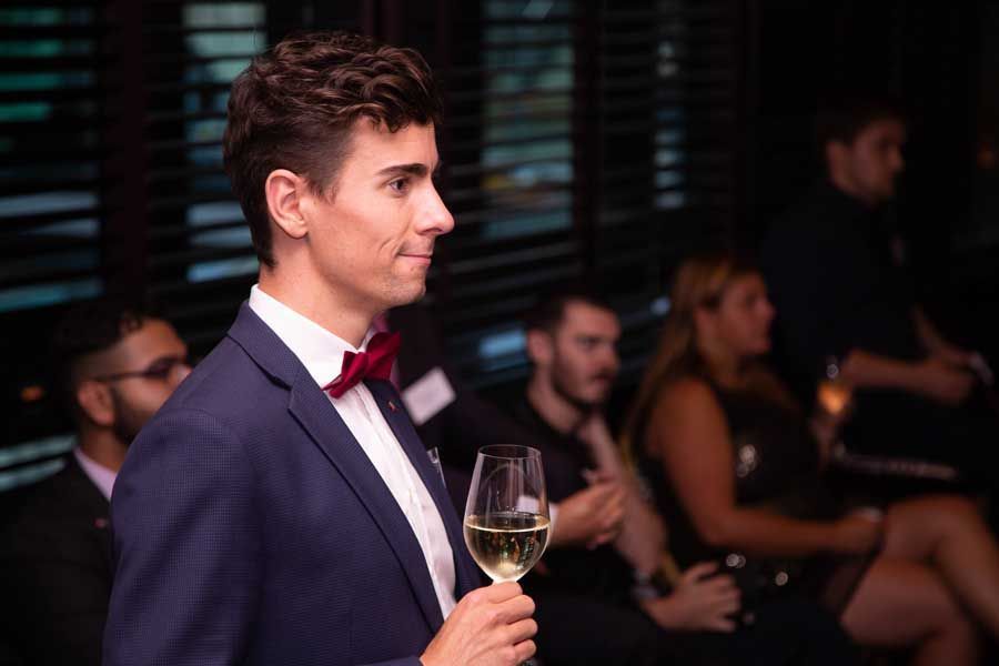 Man in blue suit and red bow tie holds a glass of champagne at an event.