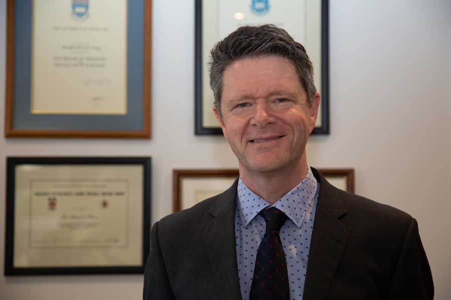 Man in suit smiles, standing in front of framed documents on a wall.