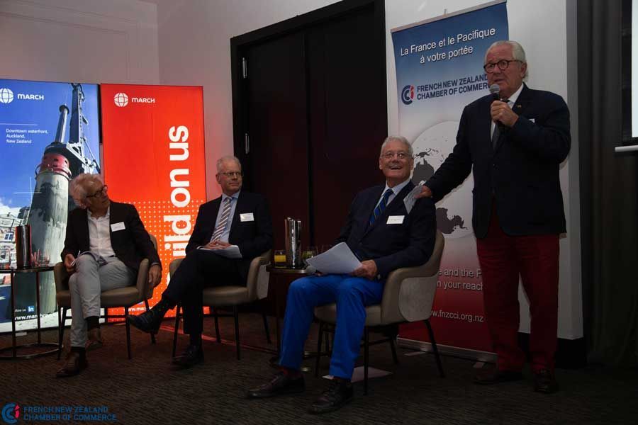 Panel of four men at a podium discussing a topic, with posters in the background.
