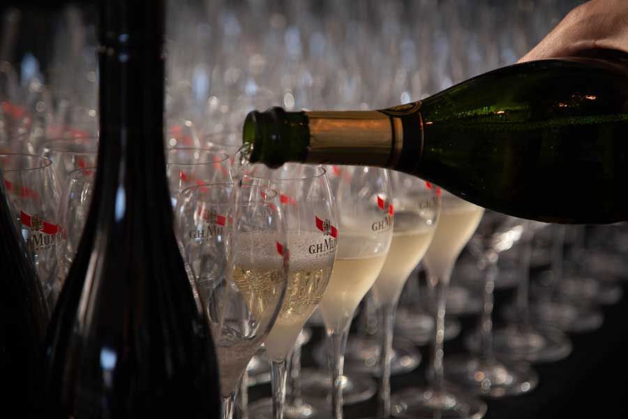 Champagne being poured into a row of glasses; bottles in the background.