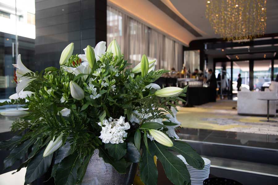 Close-up of white lilies and greenery in a vase on a table, with a blurred hotel lobby in the background.