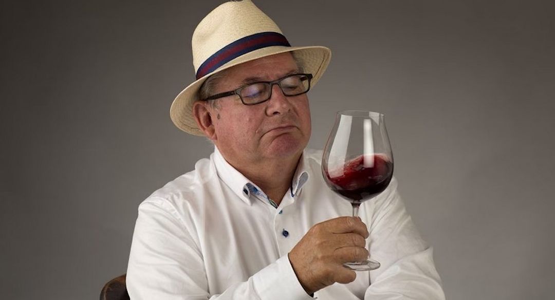 Man in straw hat and glasses swirling red wine, looking at it with a contemplative expression.