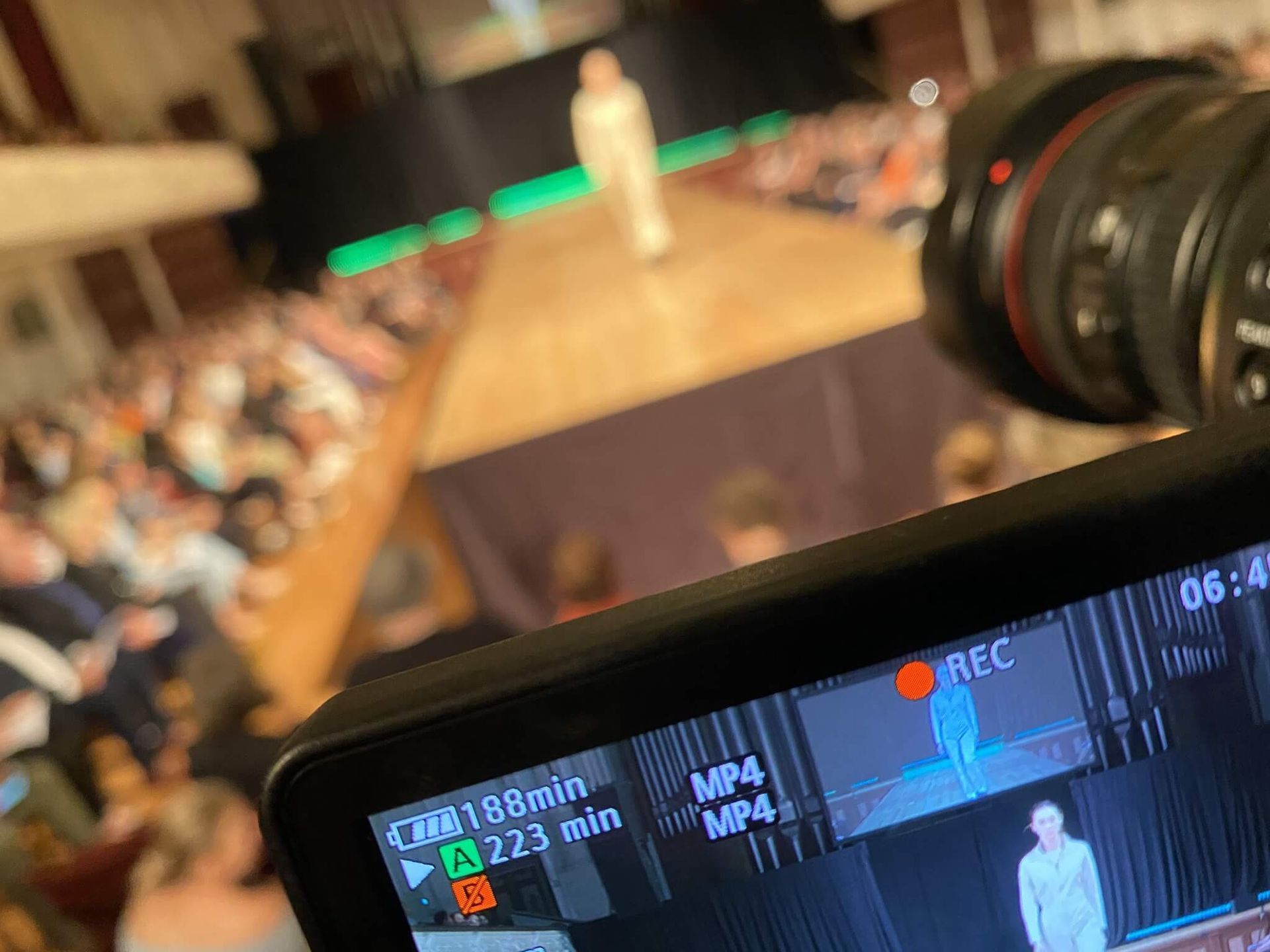 Camera filming a fashion show runway. People seated in the audience. The screen displays a live feed of a model.