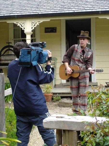 Man in costume playing guitar being filmed outdoors near a yellow building.
