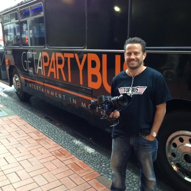 Man with video camera in front of a black party bus with orange lettering, 