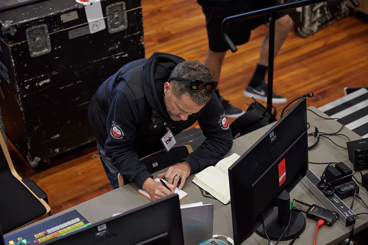 A person in a black hoodie writing at a desk, looking down. There are two computer monitor. 