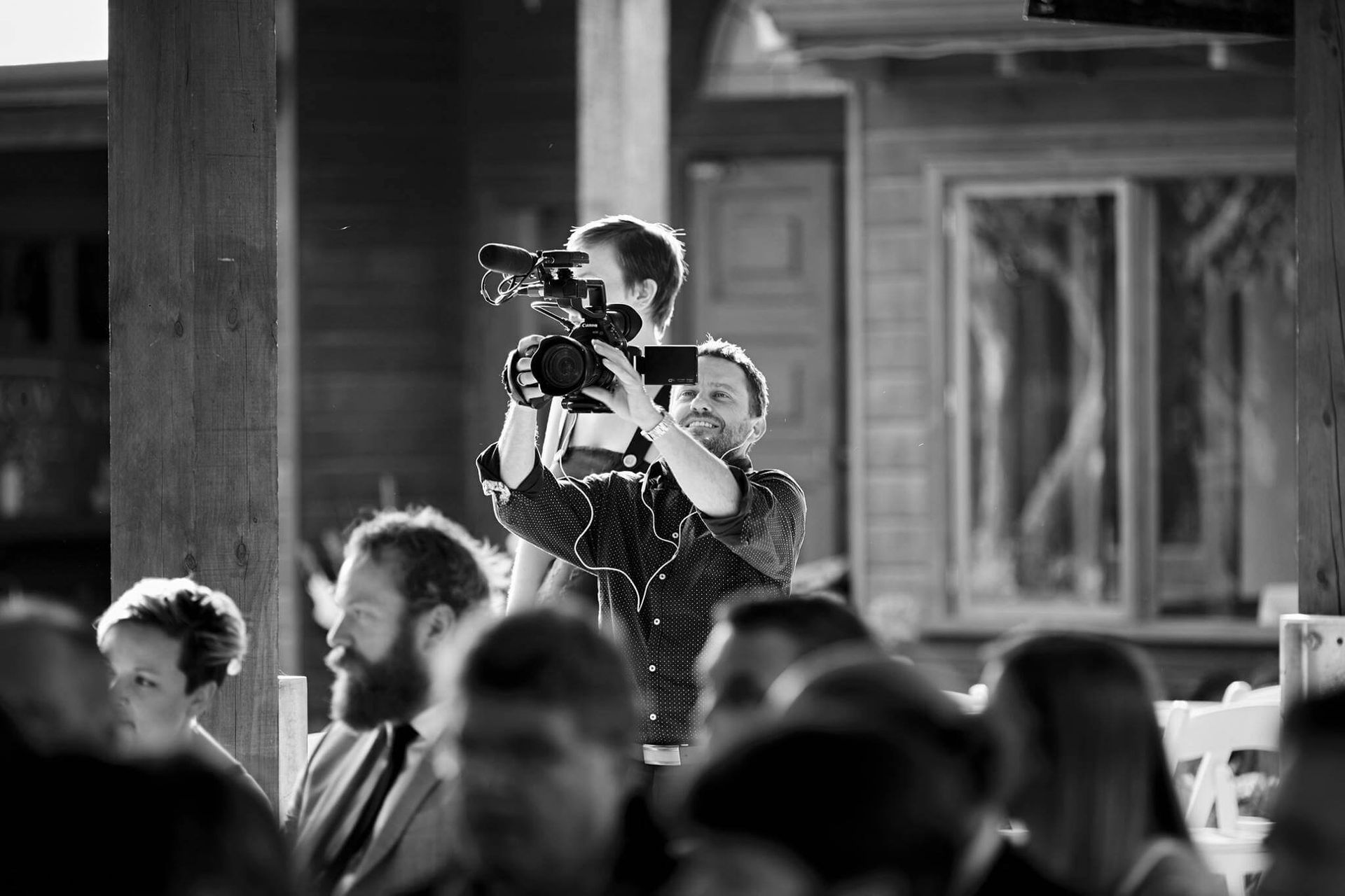 Cameraman filming event from inside wooden structure; audience seated in foreground; sunlit.