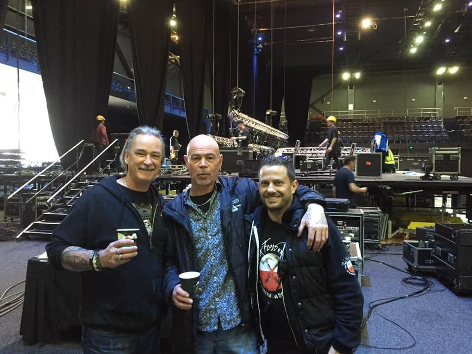 Three men pose backstage at a venue. Pink Floyd Experience  stage setup with lights and equipment visible in the background.