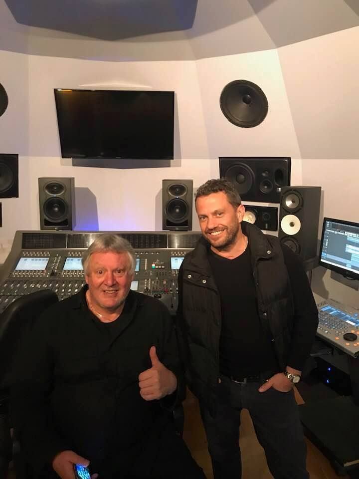 Two men in a recording studio. One gives a thumbs-up. Mixing console, speakers, and a monitor visible.