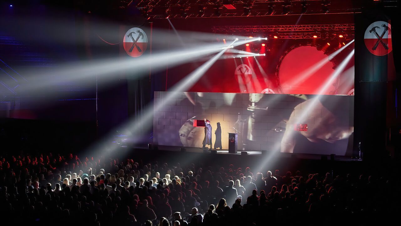 Concert stage with performers, bright spotlights, and a large screen displaying images. Audience in foreground.