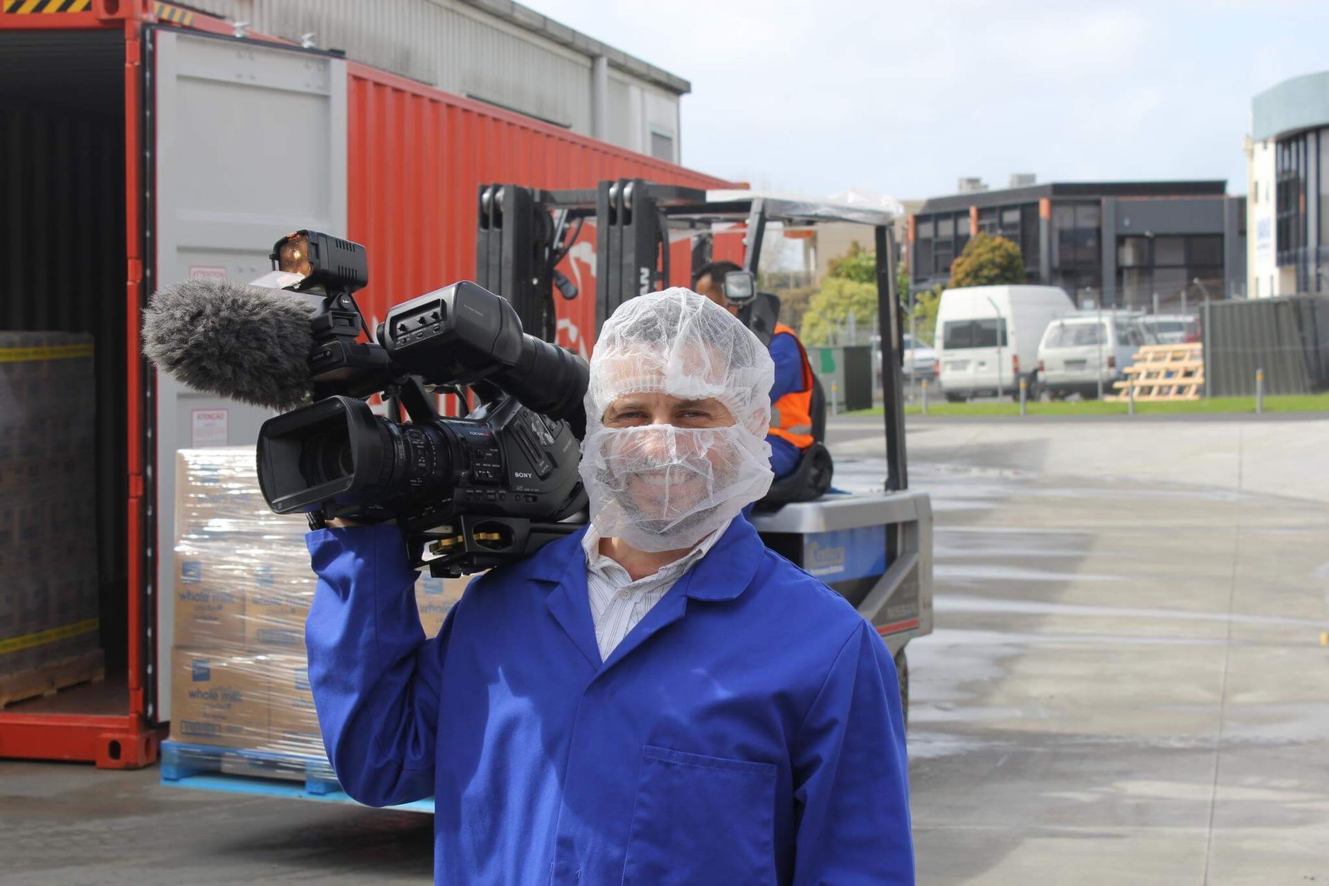 Man in blue lab coat and hairnet holds a camera, standing near a loading dock with a forklift and shipping container.