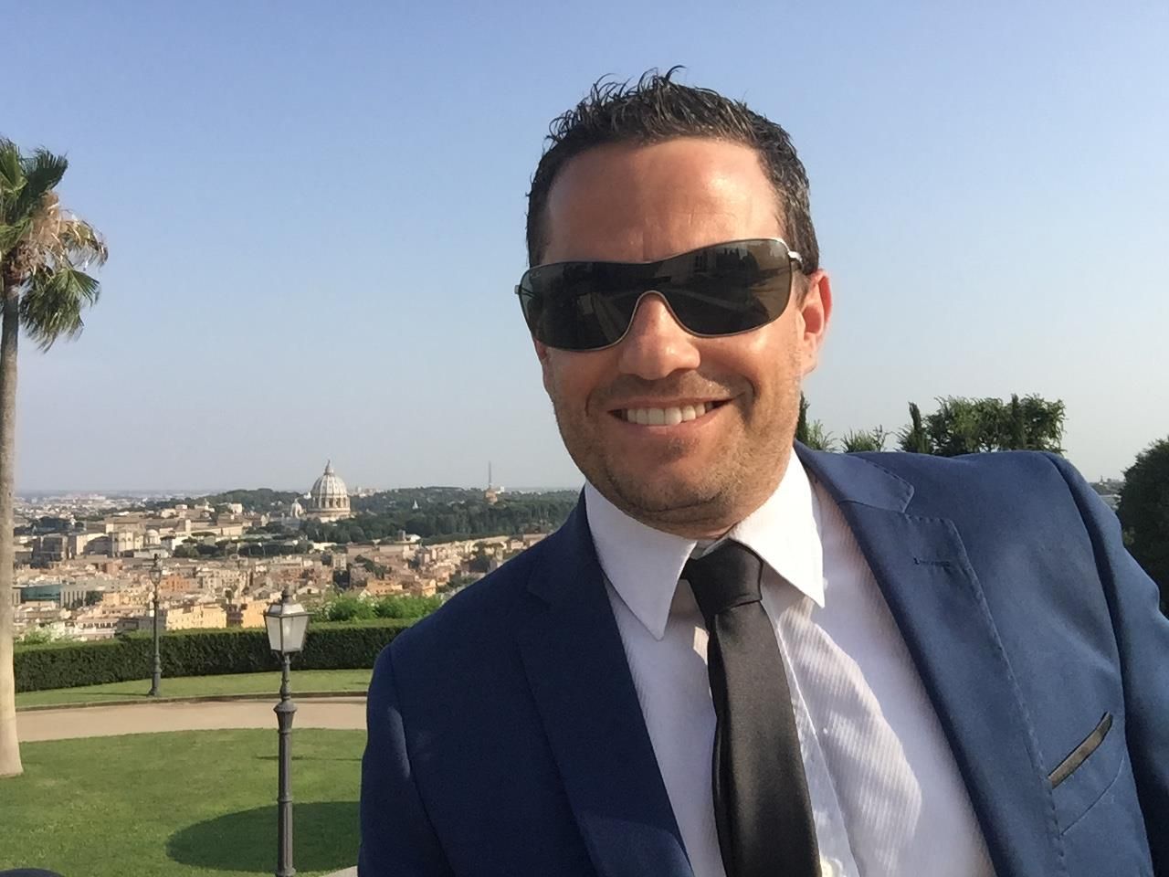 Man in blue suit and sunglasses smiles, posing with a city backdrop and green landscape.