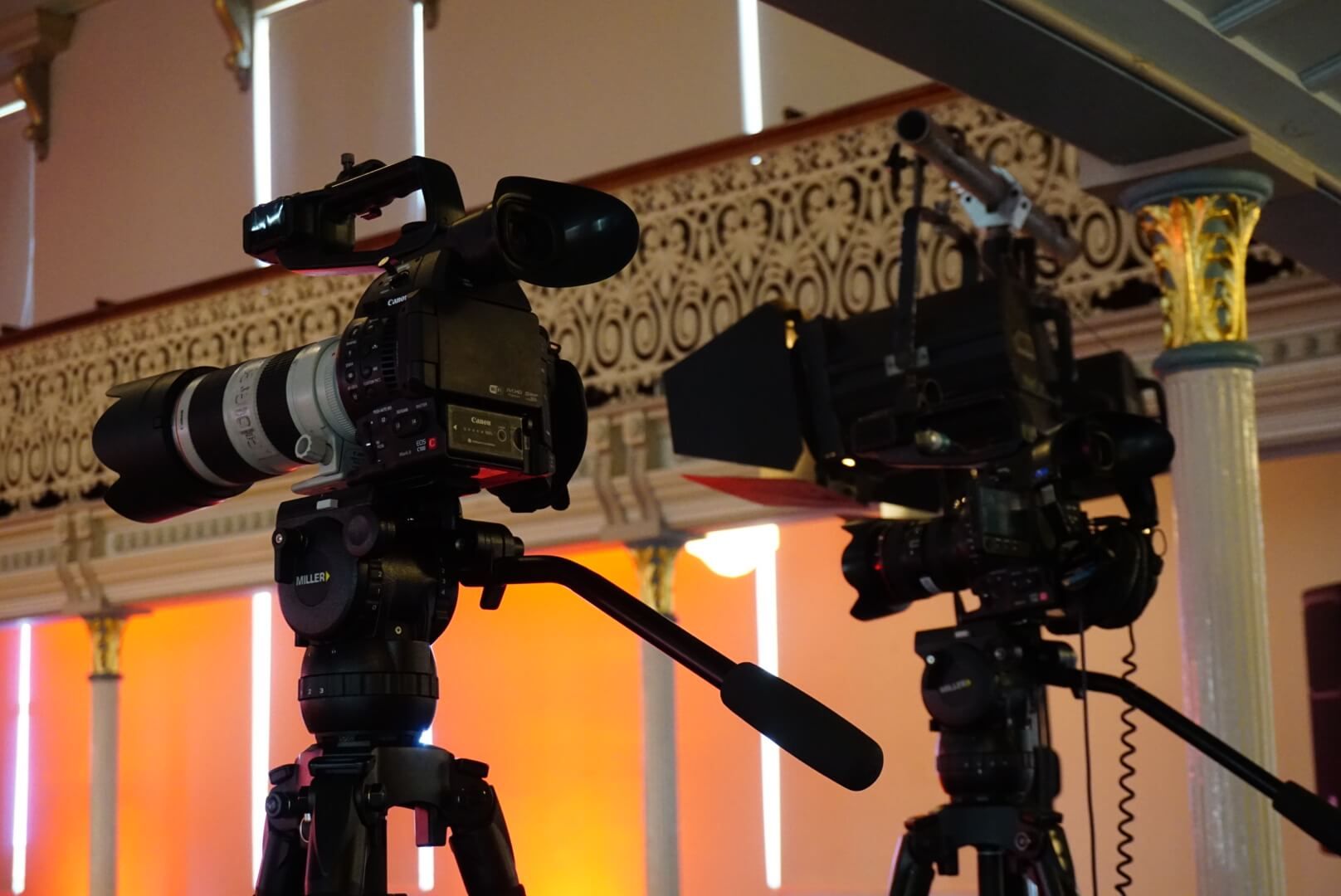 Two professional video cameras on tripods, indoors, pointed forward.
