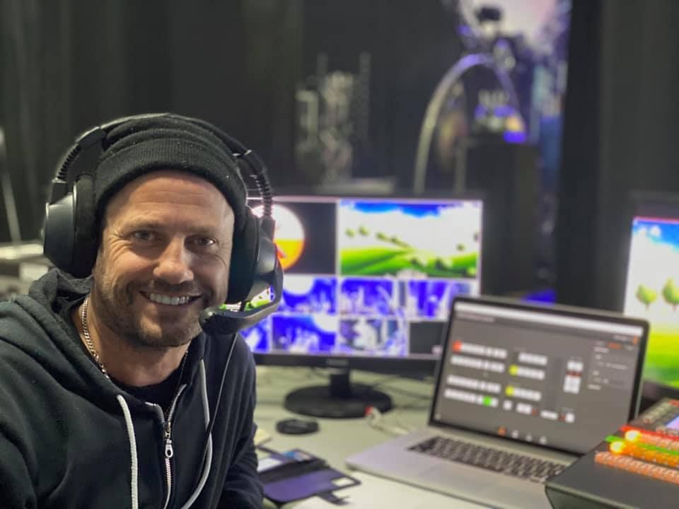 Man wearing headset in a control room, smiling at the camera, with monitors showing video feeds.
