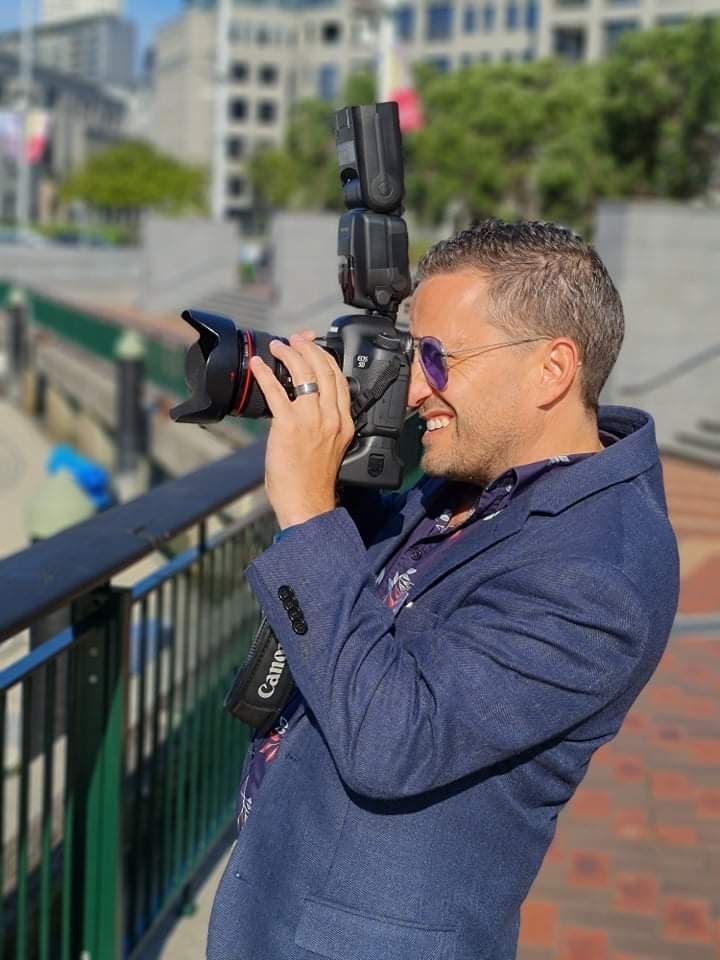 Man in a blazer and sunglasses photographing with a camera; city backdrop.