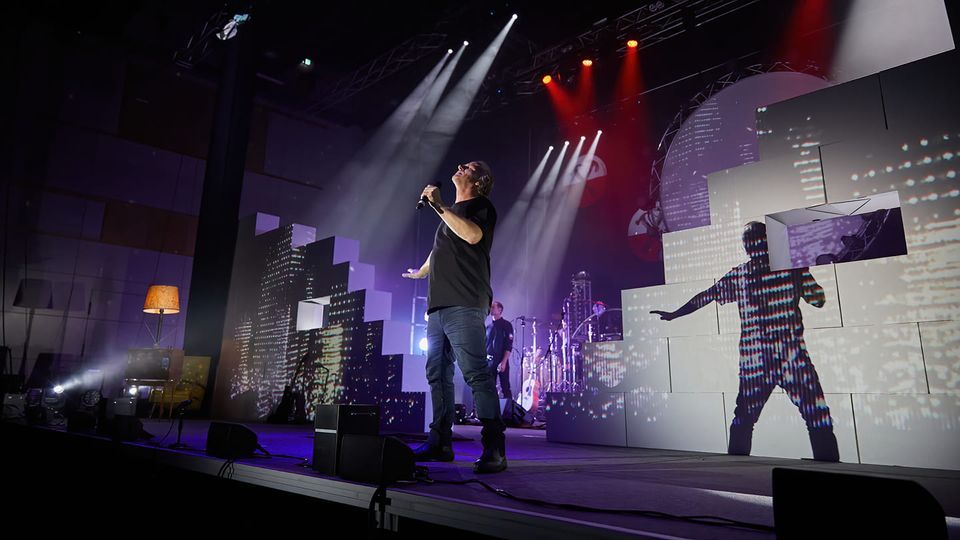 Pink Floyd Experience Singer on stage performing; city backdrop; stage lights illuminate the performer.