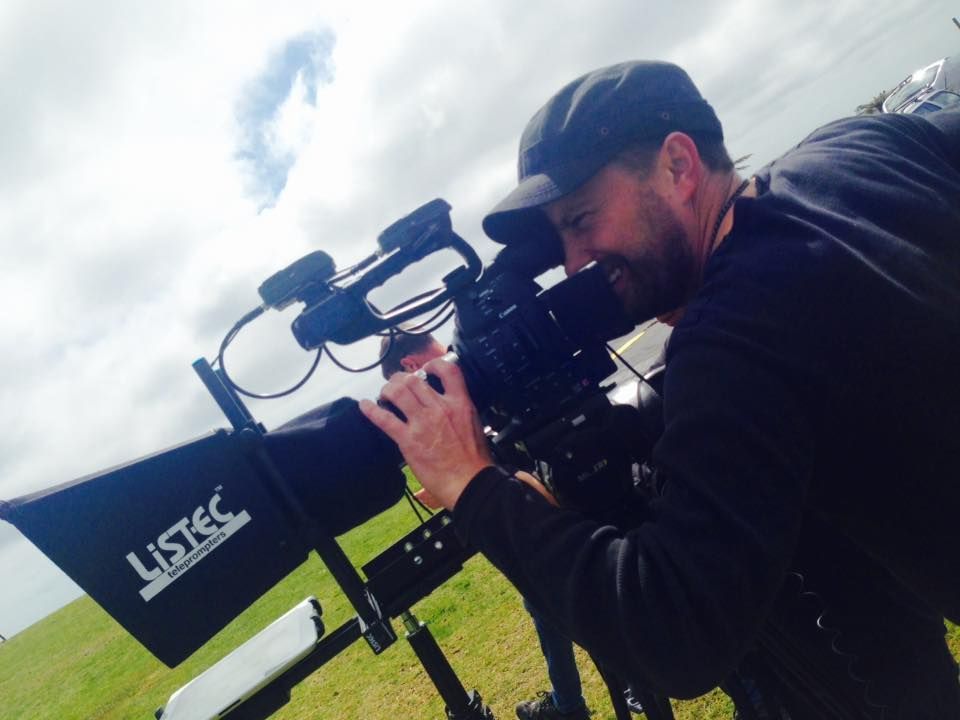 Man filming with a camera on a sunny day, smiling and wearing a black cap.