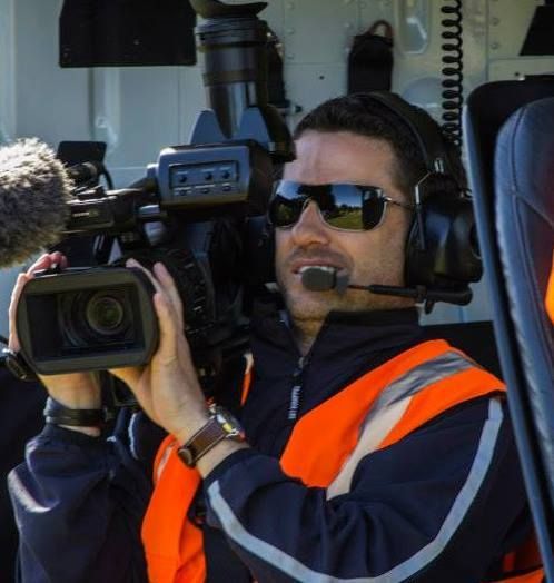 Cameraman wearing sunglasses and headset, filming outdoors. He has an orange safety vest and holds a video camera.