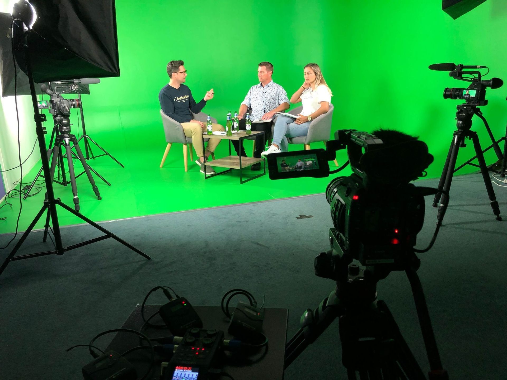 Three people in chairs, being filmed in front of a green screen, with cameras and lights.