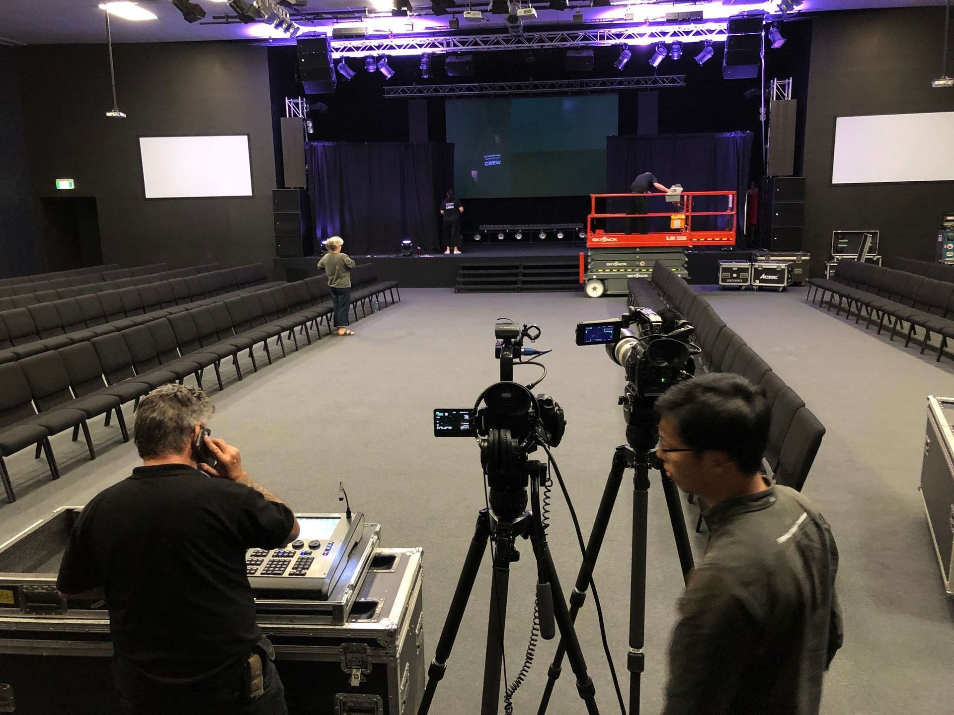 Two cameras on tripods face a stage. Crew members work to set up for an event in an auditorium.