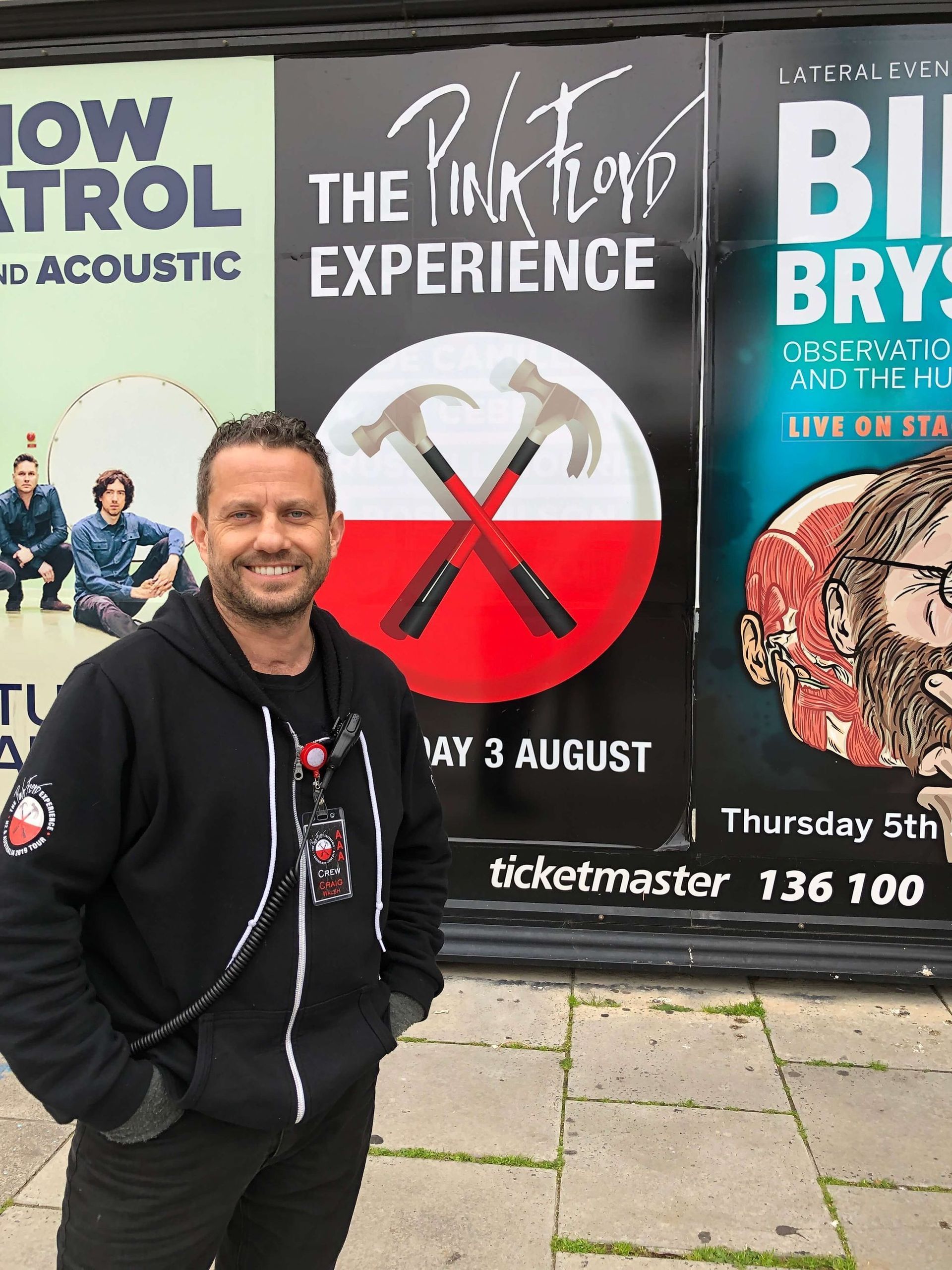 Man smiles in front of Pink Floyd Experience, Snow Patrol, and Bill Bryson posters.