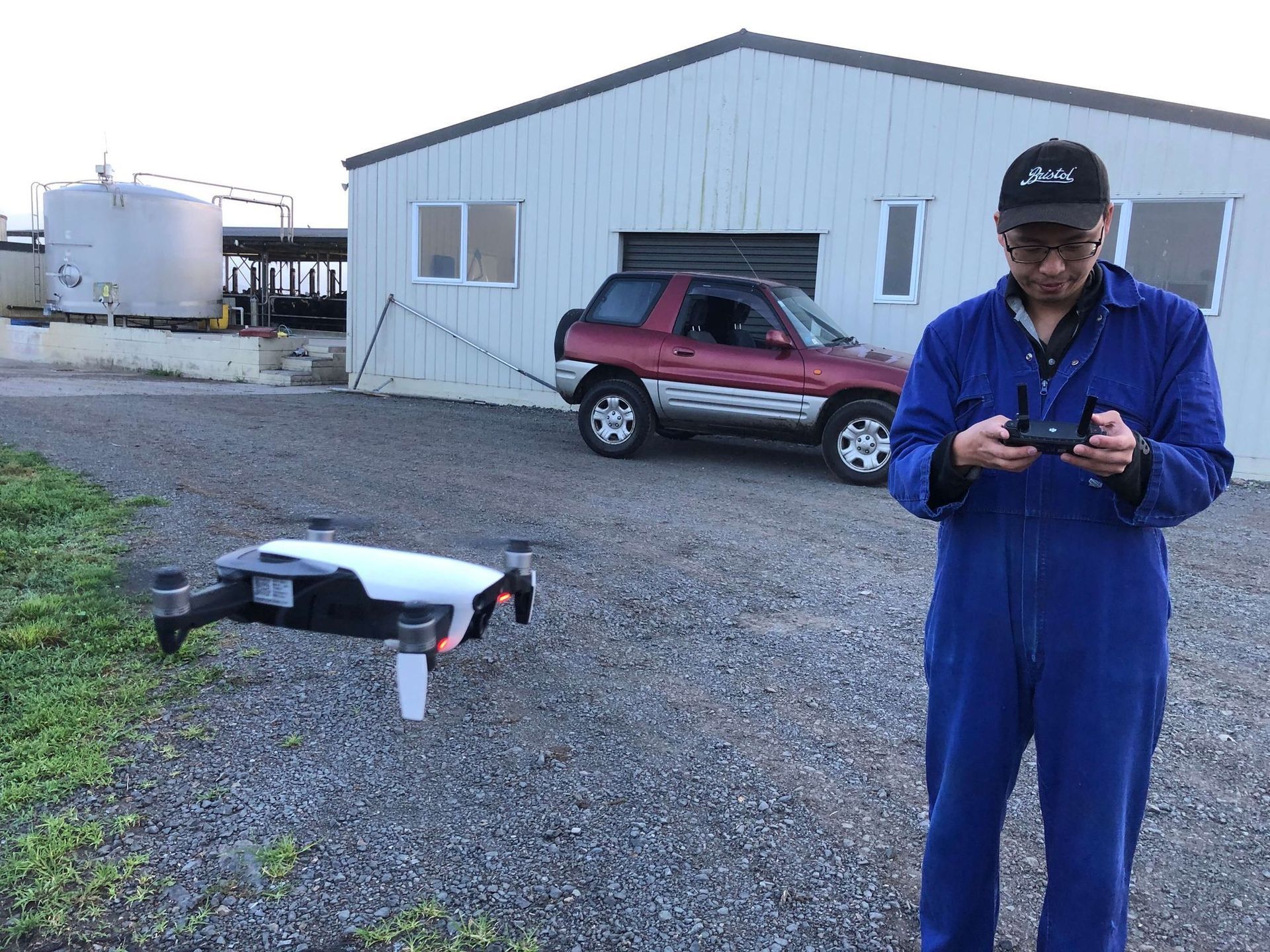 Man in blue jumpsuit operating drone on gravel with a car and building in the background.