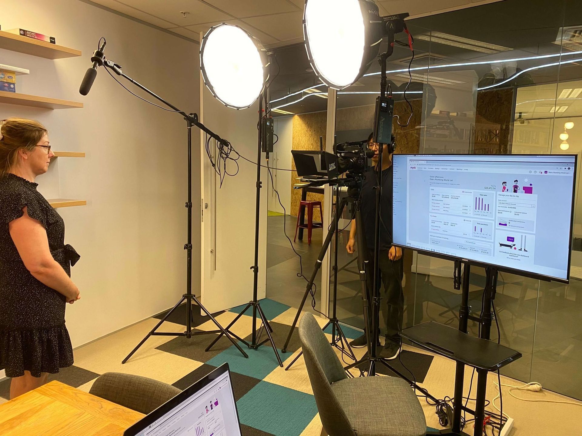 Woman being filmed in a studio with lights, microphones, a camera, and a monitor displaying graphics.