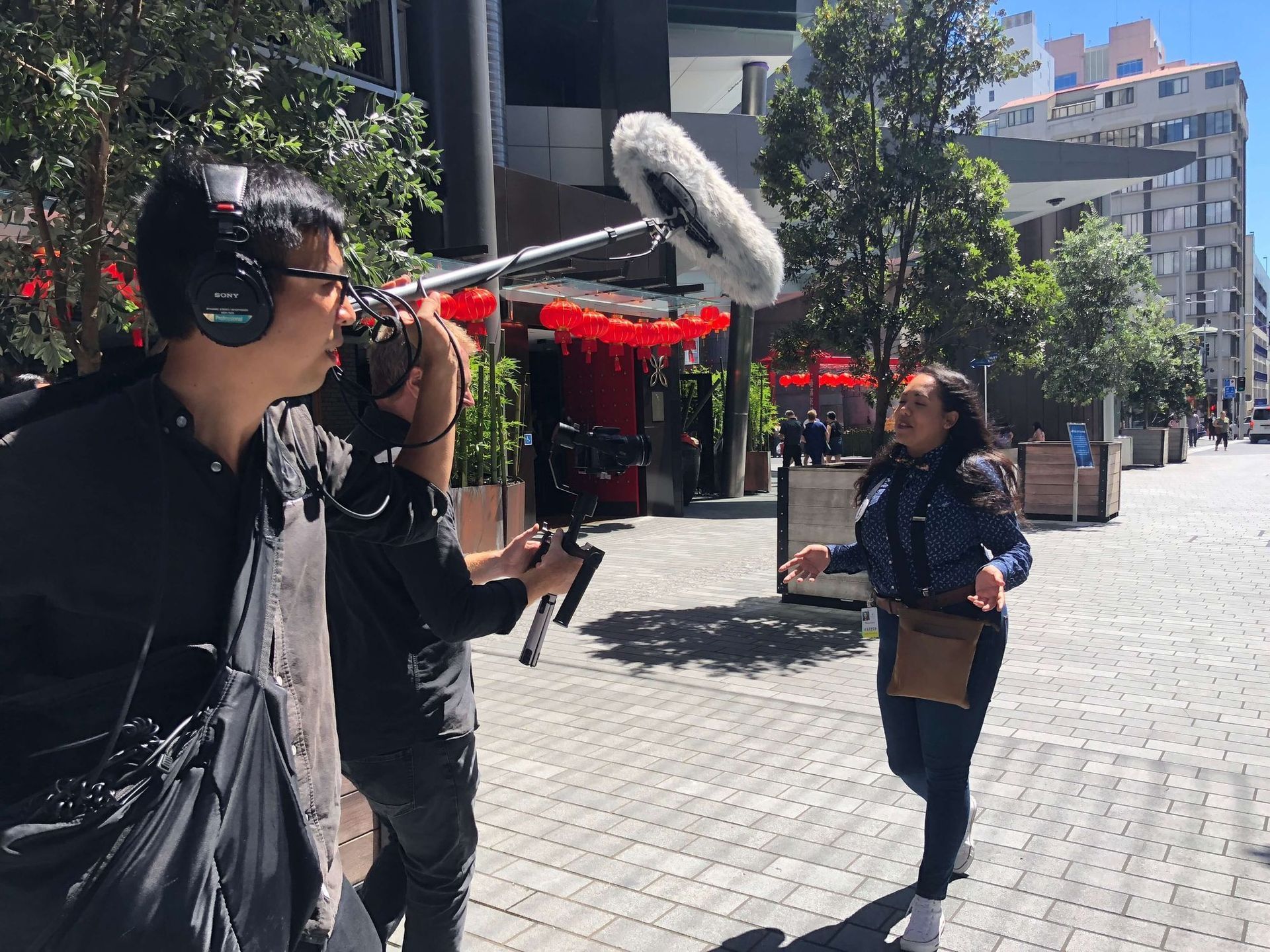 Film crew recording a woman speaking outdoors. Sound recordist, camera operator, and talent in city setting.